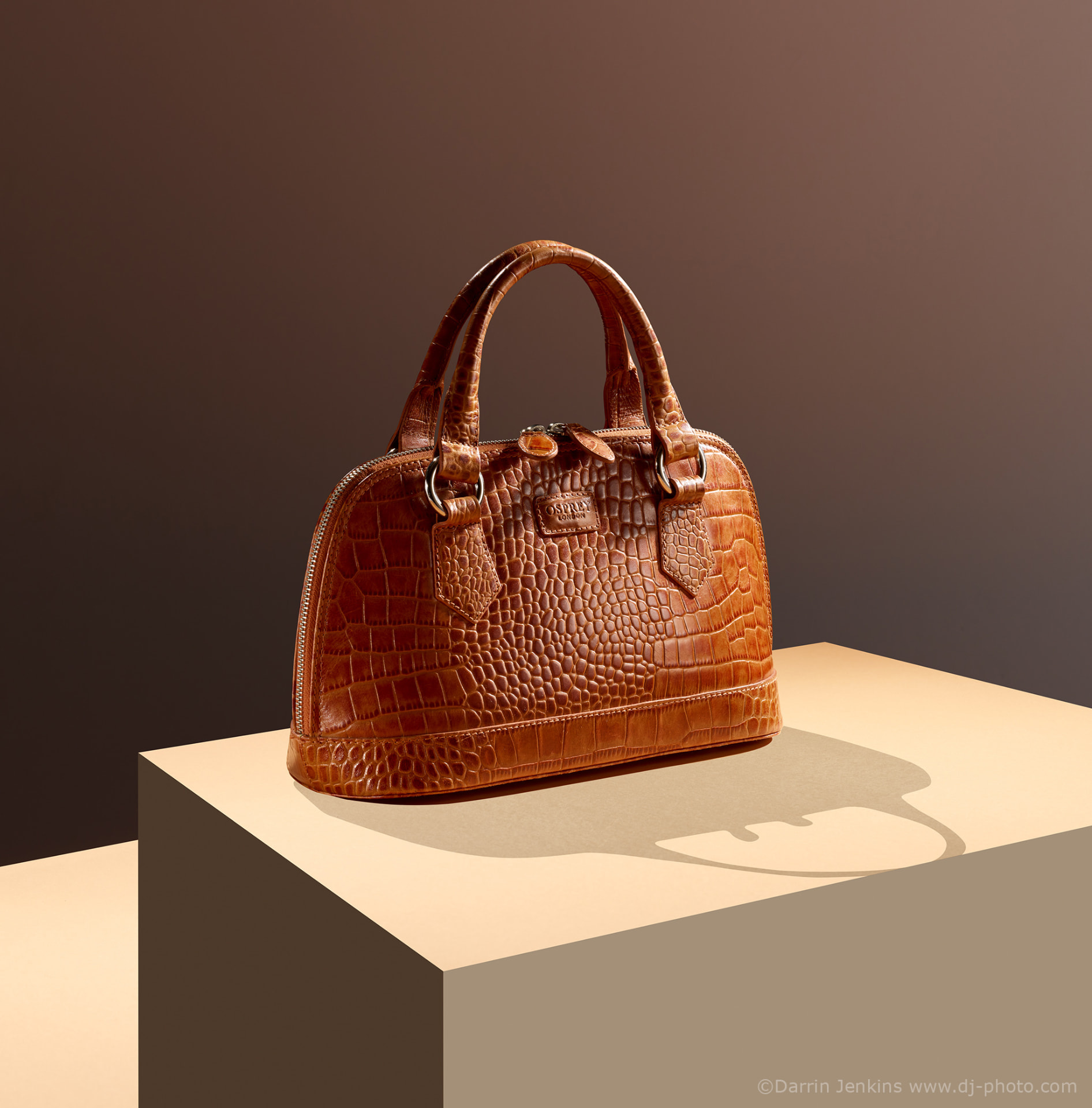 A tan crocodile-embossed leather handbag photographed on a stone plinth in studio light